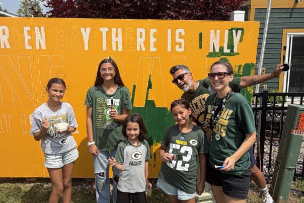 Family of 6 smiling for a picture in front of the Green Bay community fence that they were helping to paint.