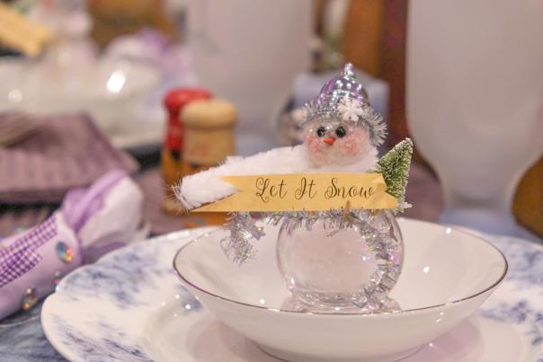sparkly snowman ornament holds tiny "let it snow" banner. it sits in a vintage place setting.