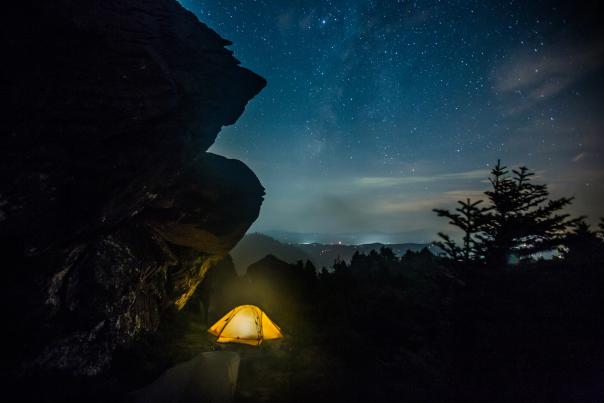 A tent placed below a rocky outcropping emits a yellow glow, with stars appearing in the nighttime sky