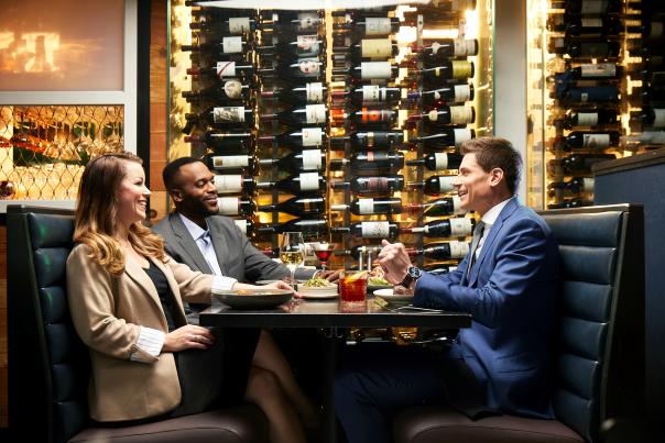 Three business leaders chat over dinner at a fancy restaurant. On the left are a white woman in a tan blazer and a black man in a gray suit. On the right is a white man in a navy suit. The table is set with steak, chicken, salad, and alcoholic drinks. A floor-to-ceiling wine display is on the wall behind them.