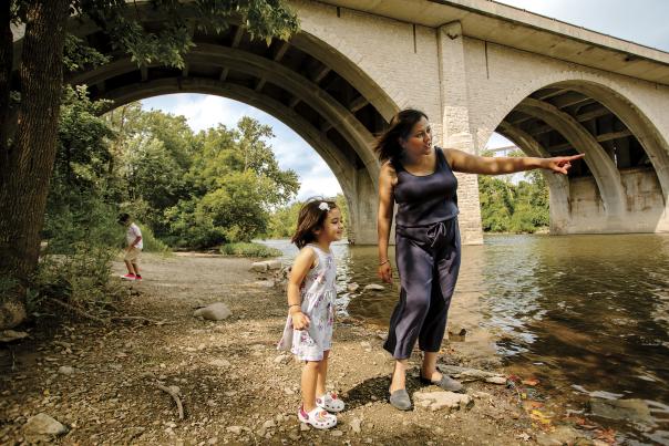 Woman and child explore the riverbank under a stone arch bridge at Dublin Springs Park on a sunny day.