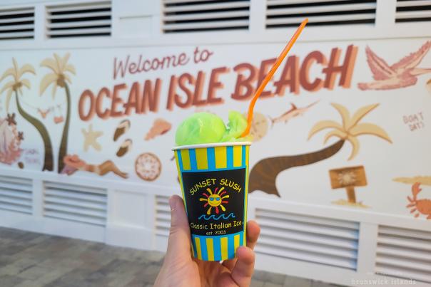 a cup of green italian ice from Sunset Slush pictured in front of a mural on Ocean Isle Beach, NC