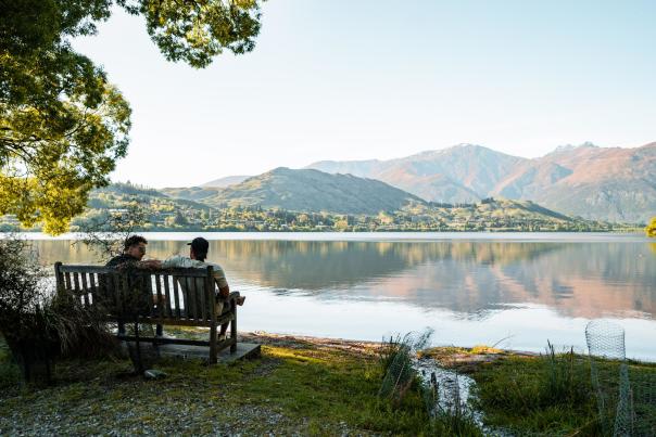 Two people sitting on a bench looking our over a lake with mountains in the background