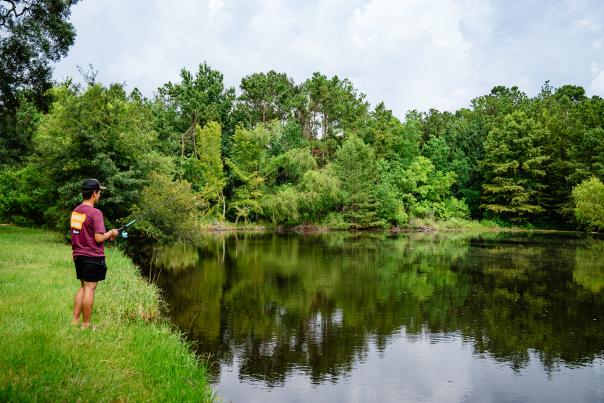 A young man (on the left of the frame) stands on the grassy shore of a fishing pond at Bear Branch Sports Fields. He is wearing a red t-shirt, black shorts, a black and tan ballcap, and he's holding a black and teal fishing pole. The water is dark and smooth. The trees on the far shore are various shades of bright green. The sky is cloudy but bright.