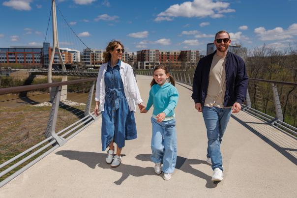 A family of three walks hand-in-hand across The Dublin Link bridge on a sunny day, with modern buildings in the background.