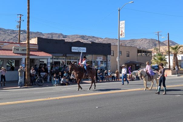 Horseback riders, including one carrying an American flag, in the 2024 Pioneer Days Parade in downtown Twentynine Palms, with sidewalk spectators, local storefronts, and desert mountains in the background.