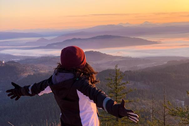A person stands with their arms spread out on a snowy peak looking out over the foggy valley with sunset colors