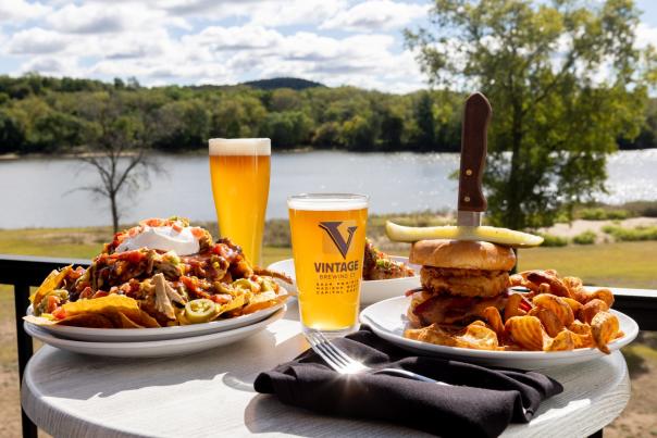 A plate of nachos, a tall cheeseburger, chips and two beers on a patio table in front of a lake at Vintage Brewing