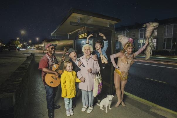 An older lady with a big beaming smile stands at a bus stopped surrounded by performers, a dancer, guitar player, dog and a little girl in a yellow coat.
