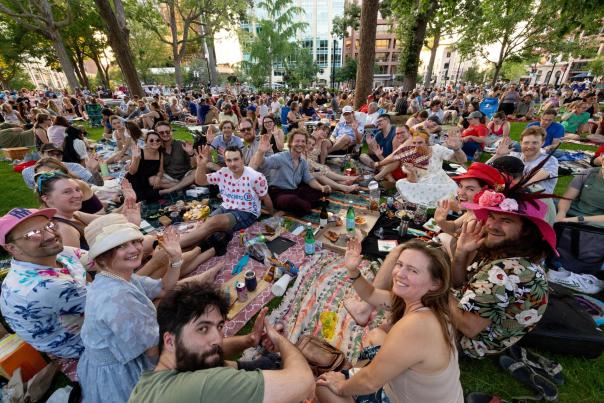 A large group of people sitting on blankets on the Capitol Square, waving at the camera.