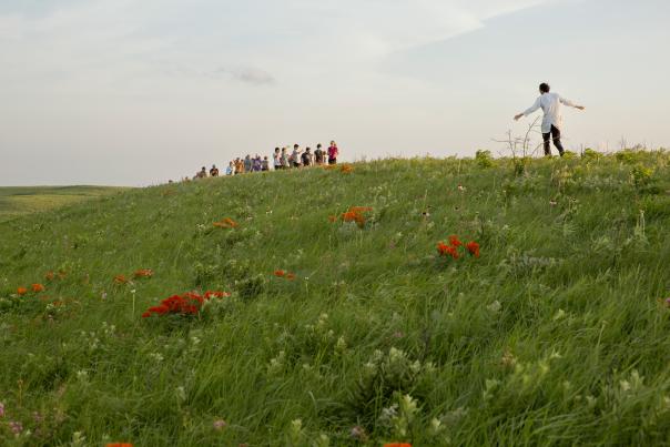 a green grass hill with orange wildflowers. People are standing on top of the hill. One man has is arms out to the side