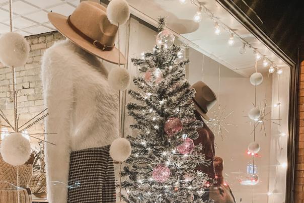 Store window display with mannequins dressed in winter clothing, a white faux fur coat, hats, a decorated Christmas tree, and hanging ornaments.