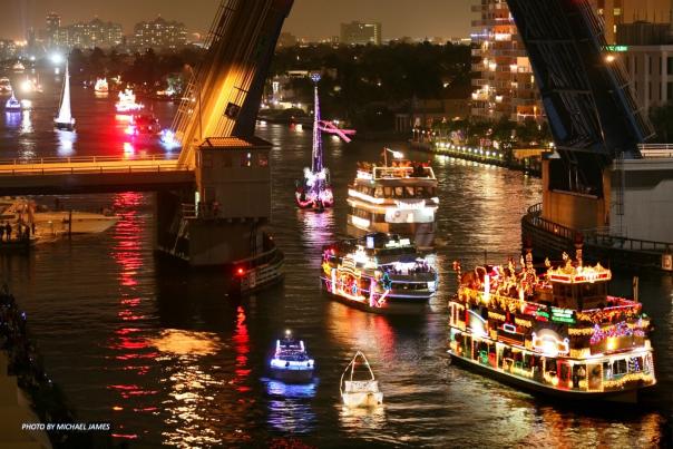 Several Boats of different sizes all lit up and decorated for the December holidays floating up the river through an open draw-bridge