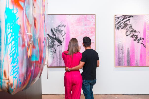 Image of a man and woman facing away from the camera looking at colorful art mounted on the white walls of an art gallery.