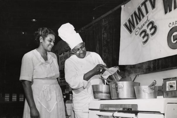 A black and white photo of a Black man, Carson Gulley, cooking on TV with a Black woman.