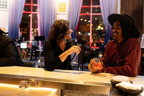 Two people sit at a stylish bar engaged in a lively conversation, each holding a drink. The woman smiles while talking to the man in a wide-brimmed hat. The setting is warmly lit with modern decor, including large windows, blue curtains, and ambient city lights in the background. Another person sits further down the bar, looking away.