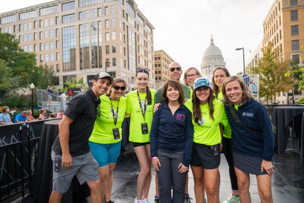 Two white men and seven white woman smile while volunteering for a shift at the IRONMAN Wisconsin finish line in downtown Madison