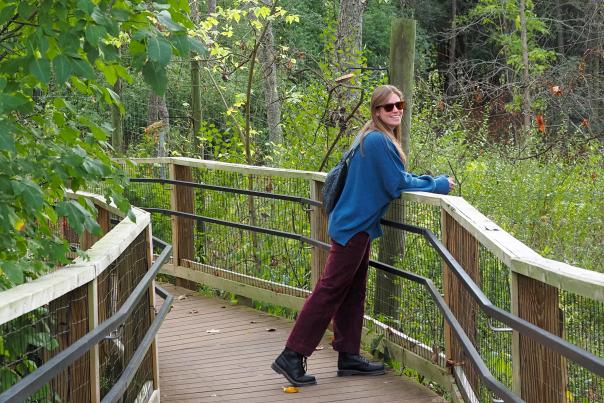 Woman leans on wooden ledge outdoors on a boardwalk surrounded by green treesh and shrubs. She turns to smile for the photo.