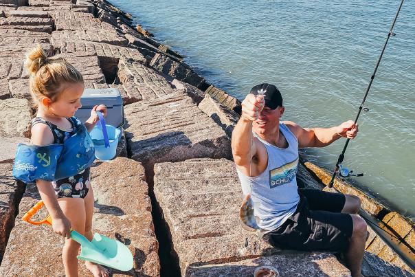 A father sitting on a jetty helps his young daughter learn how to fish.