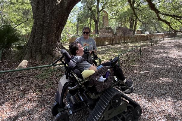 Christy Paulsell accompanies her daughter, PJ, in an all-terrain TrackChair beneath towering live oaks at Fontainebleau State Park, with the historic sugar mill ruins in the background.
