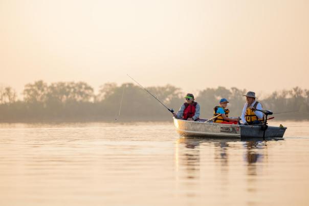 A canoe with three people paddles on calm water during sunset, surrounded by trees and a serene landscape. The scene evokes outdoor adventure.