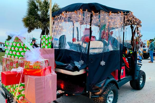A golf cart is decked out with Christmas lights. On a trailer, it pulls a bundle of Christmas gifts.
