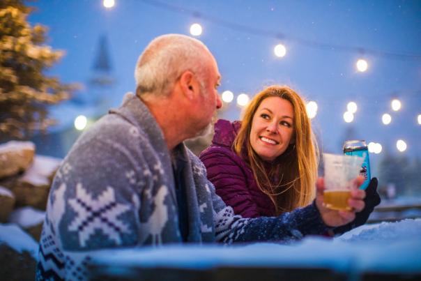A couple in sweaters hold beverages and gaze at each other with fairy lights and snow falling above them.