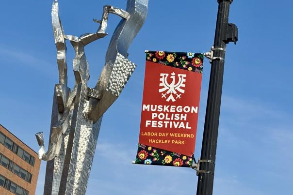 Abstract outdoor sculpture reaching towards the sky next to a streetlamp and a banner announcing the Muskegon Polish Festival at Hackley Park on Labor Day weekend.
