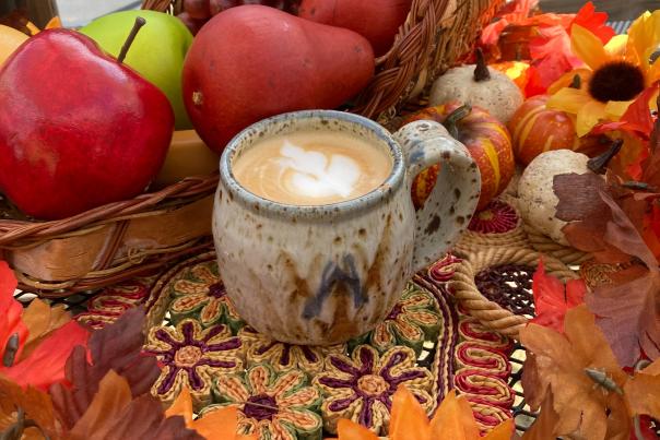 A latte sits in a handmade mug, surrounded by fall leaves and apples.