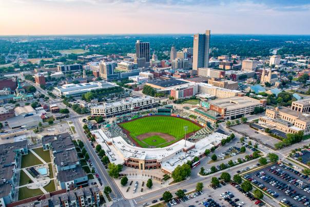 Aerial view of Parkview Field in downtown Fort Wayne during a TinCaps baseball game. Showing a vibrant cityscape featuring the baseball stadium surrounded by urban buildings, parks, and parking areas. The skyline showcases a mix of modern architecture and green spaces.
