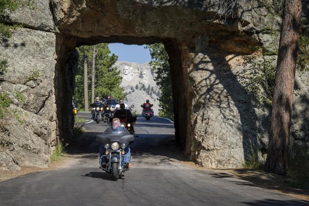 Motorcyclists ride through a rocky tunnel with Mount Rushmore in the background. The scene is set in a forested area, conveying a sense of adventure.