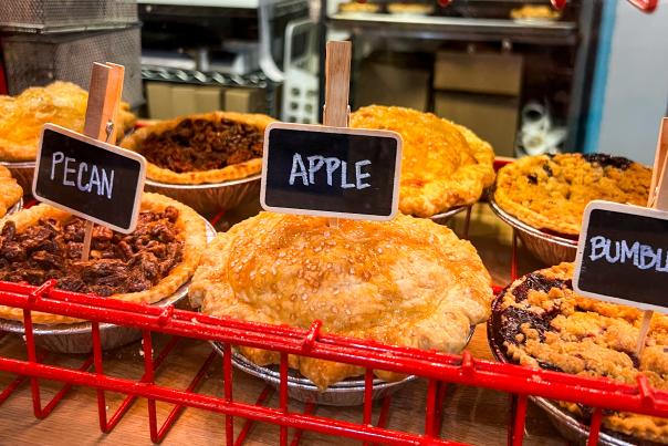 Seven pies sit atop a red bakery rack at Upper Crust with signs poked in the pies that read "pecan" and "apple".