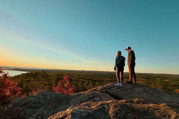 A man and a woman at sunrise during fall, standing on an a rock ledge overlooking Marquette and Lake Superior.