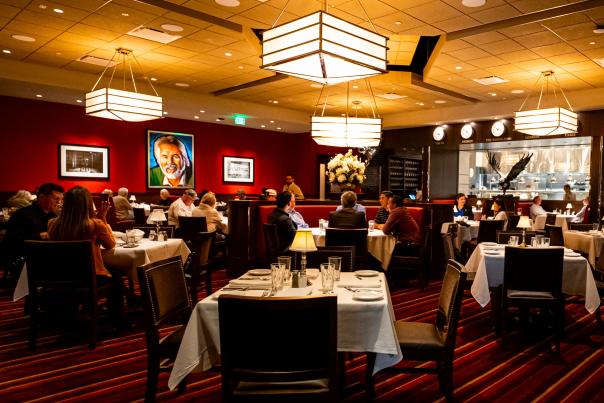 The camera overlooks a low-lit dining room with round booths clustered in the center with square tables aligned around them. People are seated at tables throughout the room, though not directly in front of the camera. There is a window looking into the kitchen in the back right with an eagle sculpture in front of it.