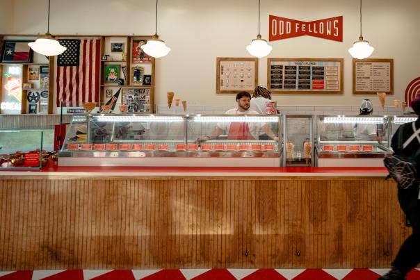 OddFellow's wood-paneled ice cream counter stretches across the frame. The floor tiles are diamond-shaped, alternating in red and white. The wall behind the counter features the OddFellow's logo/banner, a menu board, and a corkboard lined with an American flag and vintage sports memoriobilia.