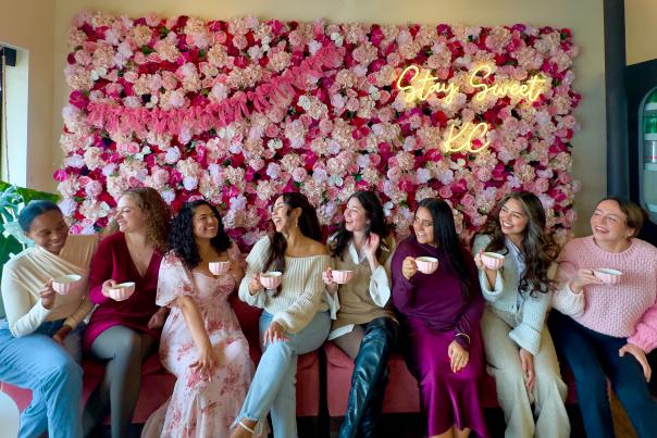 Group of 8 girls with tea cups laughing in front of the pink rose wall at Kate Smith Soiree.