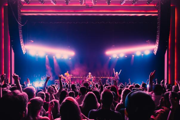 Crowd at a concert at Channel 24, with a band performing on stage under bright pink and white lights as audience members raise their hands and phones.