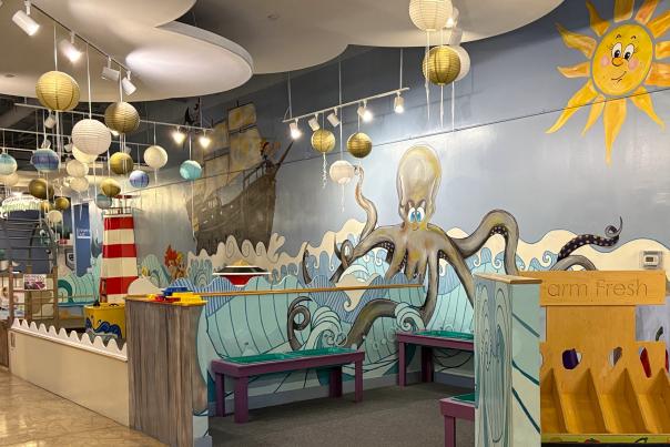 The camera focuses on a play area within The Woodlands Children's Museum. The walls are painted with cartoon-like waves, a pirate ship, a giant octopus, and a smiling sun. In the back, there is a red-and-white striped lighthouse. On the right, there is part of a farmer's market exhibit visible.