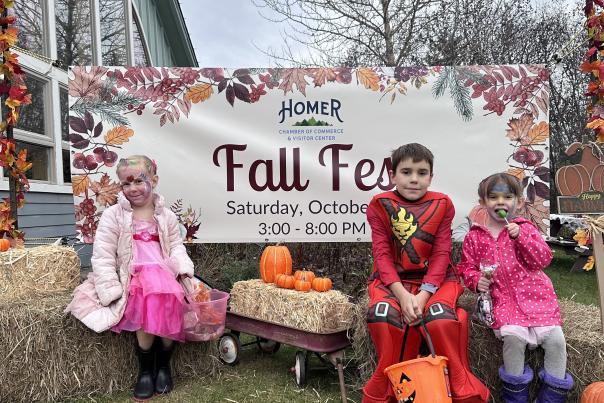 Three children dressed in colorful costumes sit on hay bales in front of a "Fall Fest" banner at the Homer Chamber of Commerce & Visitor Center. The banner announces the Fall Fest event, held on Saturday, October 26, from 3:00 to 8:00 PM. The kids are surrounded by small pumpkins and autumn decorations, embodying the festive spirit of the event.