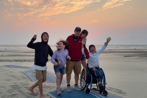 A family celebrates with hands in the air as they use a Mobi-mat all together to cross a flat sandy beach.
