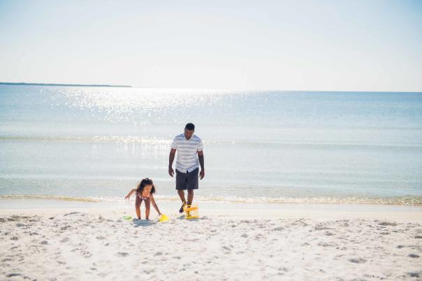 man watching daughter play with sand toys on the beach