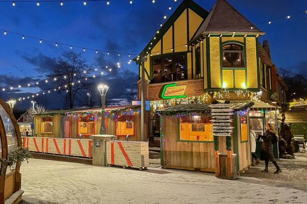 Outdoor dining area in the Village at dusk with string lights, a wooden building decorated for winter, and a transparent igloo-style dome with people inside on a snowy ground.