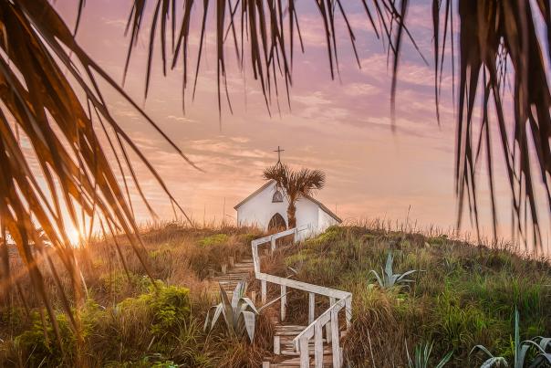 On top of a grassy hill at sunset sits a small white chapel topped with a cross. Steps with a white railing lead up to the chapel and palm tree fronds frame the photo