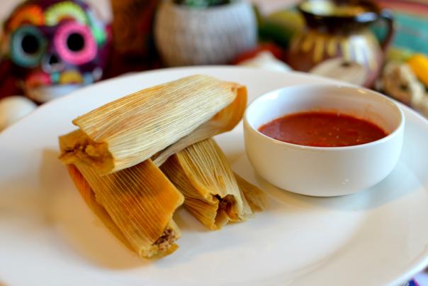 3 tamales in corn husks sit on white plate next to white bows filled with red sauce