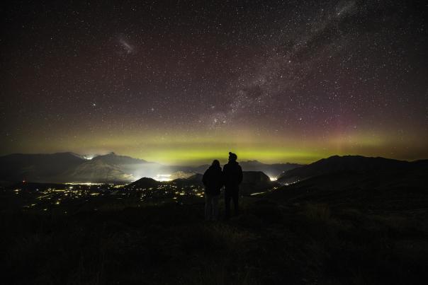 People standing on a mountain top with a starry night sky and aroura before them