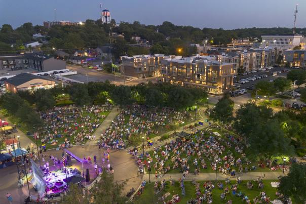 Aerial evening view of a large outdoor crowd gathered in a downtown Bowling Green park, with a lit concert stage, surrounding buildings, tree-lined paths, and city lights in the background.