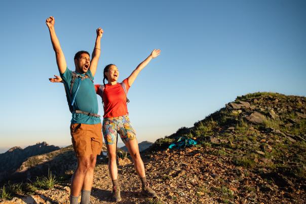 Man and Woman wearing hiking gear with their arms up and looking excited to reach the top of a mountain