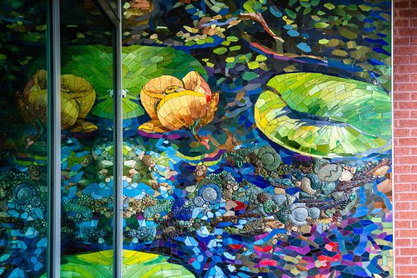 Reflections to Mist Mosaic in Stairwell at Sam Houston State University - The Woodlands. The full art piece is 4 stories tall, wrapping around the interior and exterior of the building. This segment (ground floor) is a close-up of bright blue water, bright green lily pads, bright yellow flowers, and 3D coral- or fossil-like accents.