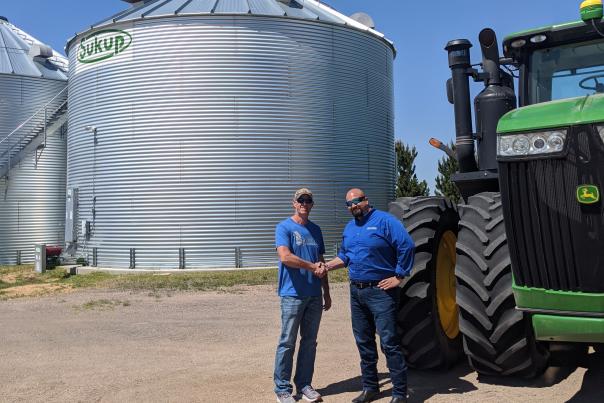 Visit Cheyenne CEO Domenic Bravo shakes hands with Rabou Farms owner Ron Rabou in front of a John Deere Tractor and grain bins on a sunny day
