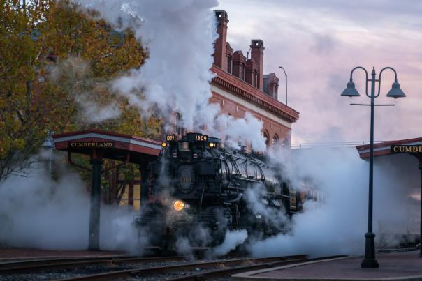 A large black steam train engine pulls into the Cumberland Station as it releases a billowy cloud of steam.